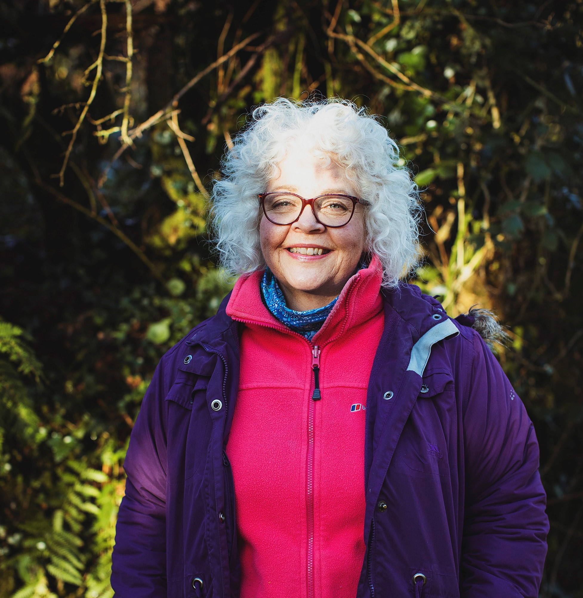 A colour photo of Chdel, a middle aged, smiling woman, with blond culy hair, glasses, and wearing out door clothes. She is standing in a wood in a path of sunlight.